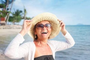 A smiling older woman on the beach wearing a straw had and sunglases